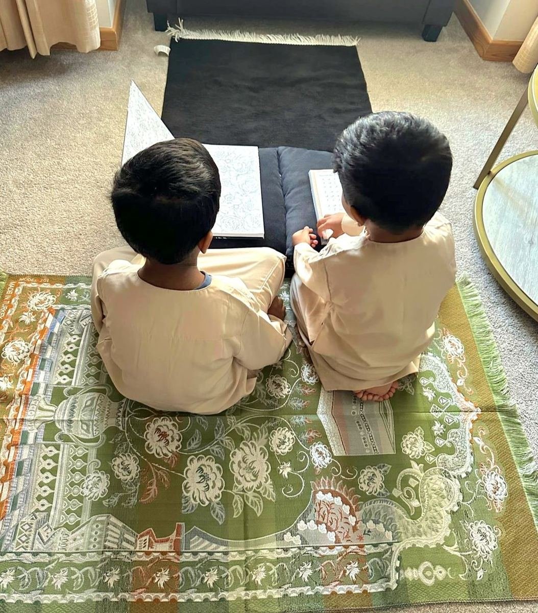 Two boys sitting side by side on prayer mats, reading.