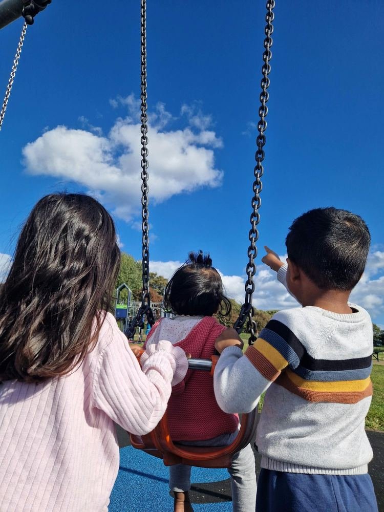 Three children seen from behind at a playground swing, one pointing up at the sky.